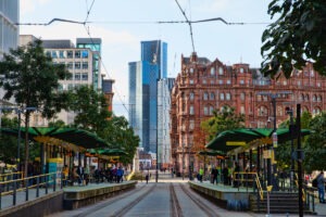 Tram station view with tracks leading into the distance. On either side, platforms shelter passengers eager for unmissable experiences. Tall modern buildings and a red-brick historic structure, hallmark Manchester attractions, stand in the background. Green trees line the street under a partly cloudy sky.